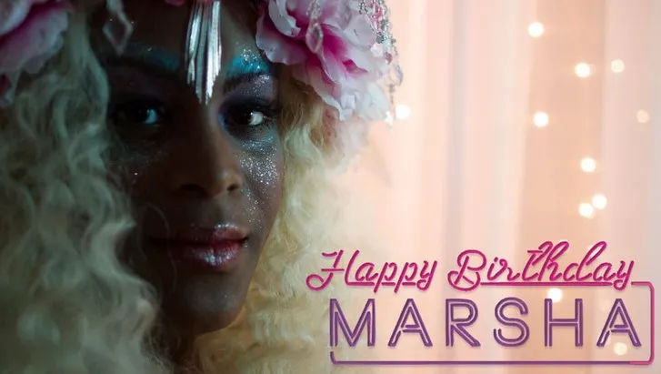 Poster for the film "Happy Birthday, Marsha!" featuring lead actress Mya Taylor. She is dressed up with flowers in her hair and sparkly glitter on her face and lips, and she's looking at the camera. The name of the film is stylized to look like a neon pink sign.
