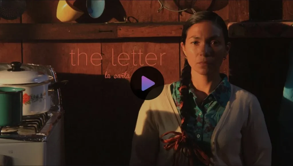 Screenshot from the short film "La carta | The Letter" of the character Rosalía (played by Myriam Bravo) sitting on a chair against the wall in her house. She is wearing a blue, patterned button-down and a white cardigan, and her long black hair is braided with a red ribbon r fabric. The expression on her face is very serious. She is sitting in her kitchen against the wall; behind her you can see cups and pots hanging, and next to her is her range with saucepans sitting on it.