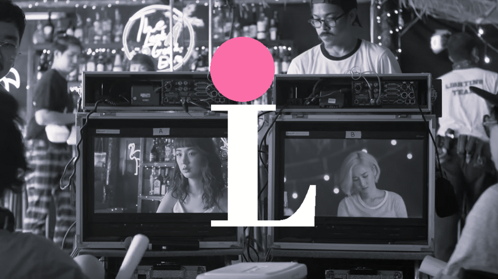 Black and white screenshot showing behind the scenes on a film set. There are two monitors labeled A and B that are showing the protagonists – on A, the lead actress (“Thanaerng” Kanyawee Songmuang) acting as a bartender, with drinks behind her, and on camera B, the second assistant named i (Sirada Tanapeng), sitting at the bar and looking down at the script. On the edges of the screenshot you can see other crew members both looking at the monitors and helping set up the scene. Superimposed on the image is a big white capital L with a pink circle on top, like the dot on top of a lower-case i