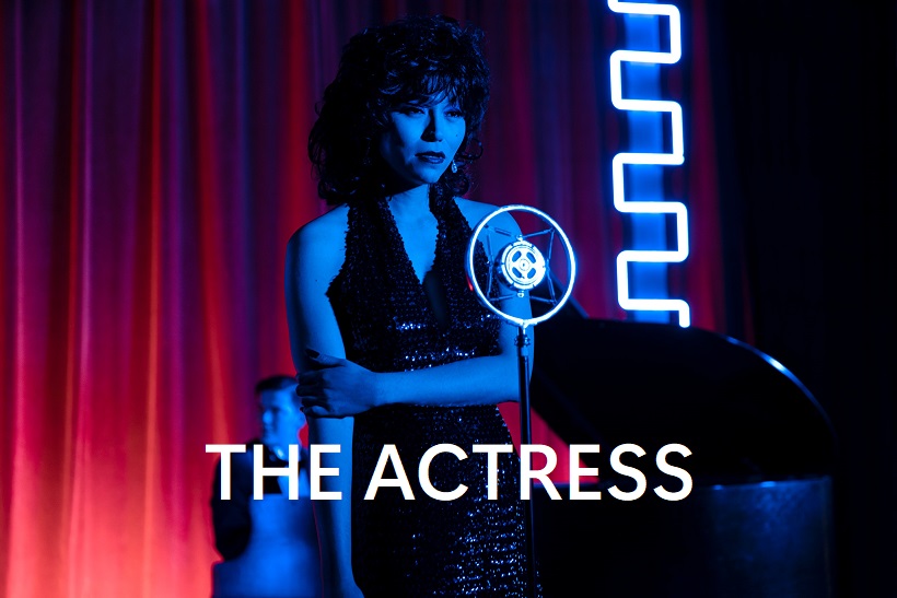 screenshot from the short film “The Actress”, starring Isabel Sandoval. Sandoval is wearing a black cocktail dress standing in front of a microphone on a stage, with a grand piano and piano player behind her, and behind them, deep red curtains. The shot, and Sandoval’s costume, hair, and makeup, are styled to make her look like Isabella Rossellini in the 1955 film Blue Velvet.