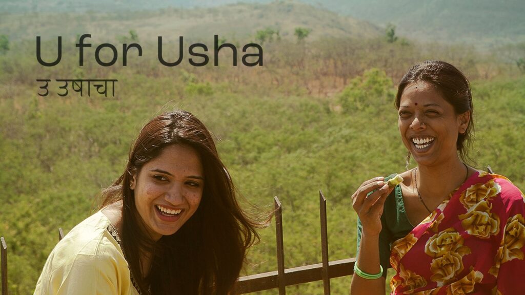 Usha (Kiran Khoje) and Manisha (Arpita Ghogardare) are both looking offscreen and laughing. Usha is wearing a sari with a green blouse and a red field with yellow flowers on it, with her hair tied up in a bun. Manisha’s long hair is loose, and she is wearing a light yellow top. Behind them are green fields.