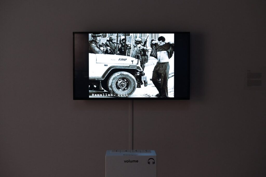 An art museum exhibit with a TV screen showing a black-and-white photo with the label “Ramallah, 2001”. The photo is of a Palestinian man faced by several Israeli soldiers and an Israeli army vehicle; the Palestinian man is lifting up his shirt to show his chest.