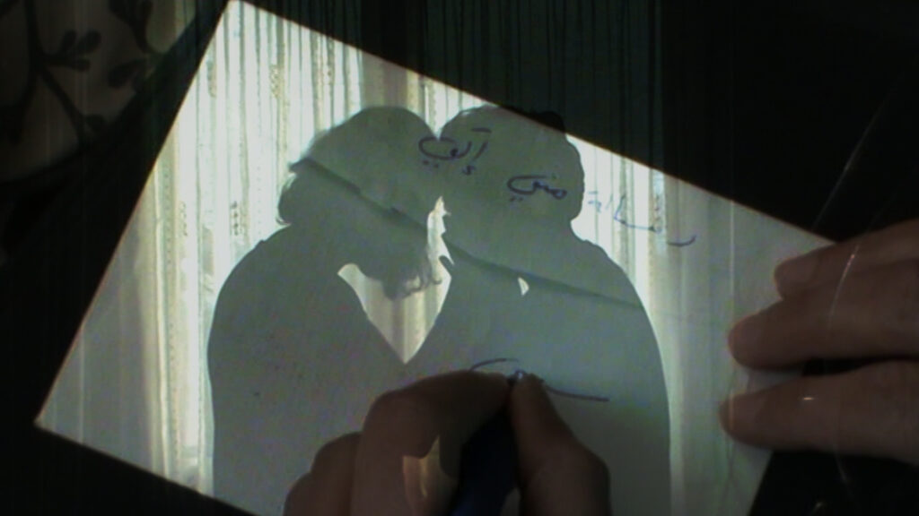 Screenshot that blends between two images: one is of a hand writing in Arabic on a piece of paper, and the other is of two silhouettes of people backlight by sun from a window behind them, embracing and touching their foreheads together.