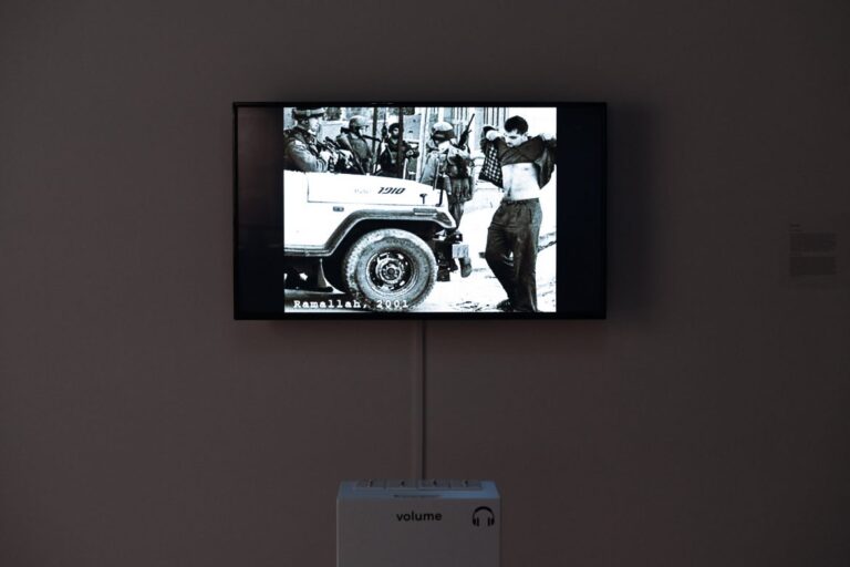 An art museum exhibit with a TV screen showing a black-and-white photo with the label “Ramallah, 2001”. The photo is of a Palestinian man faced by several Israeli soldiers and an Israeli army vehicle; the Palestinian man is lifting up his shirt to show his chest.