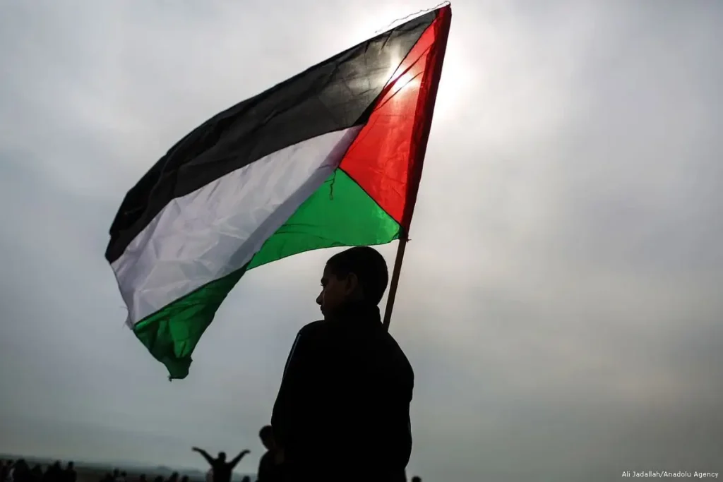 A person holds a Palestinian flag beneath a gray sky.