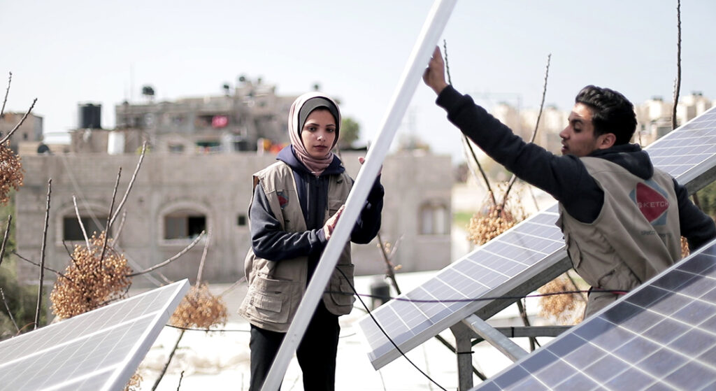 Two people, both wearing vests as technician uniforms, help place solar panels on stands.