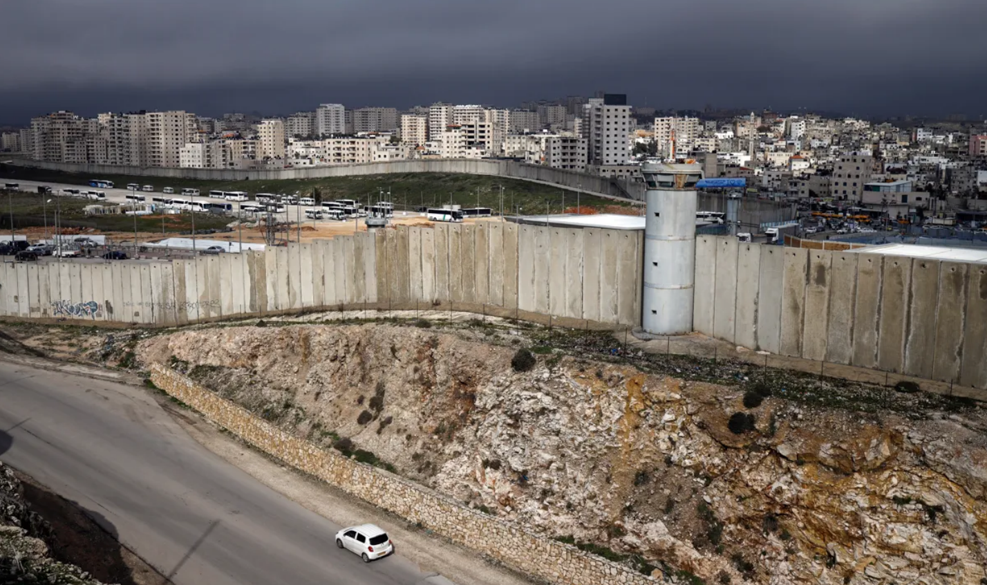 A huge concrete wall cuts through the middle of the photo; on the side closer to the camera is a road with a car driving, while on the other side are dozens of buildings and roads showing a city.