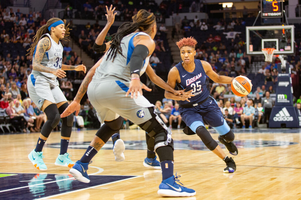 Angel McCoughtry, wearing a dark blue Atlanta Dream jersey, is moving with the ball while two Minnesota Lynx players, Erlana Larkins and Seimone Augustus, attempt to guard her