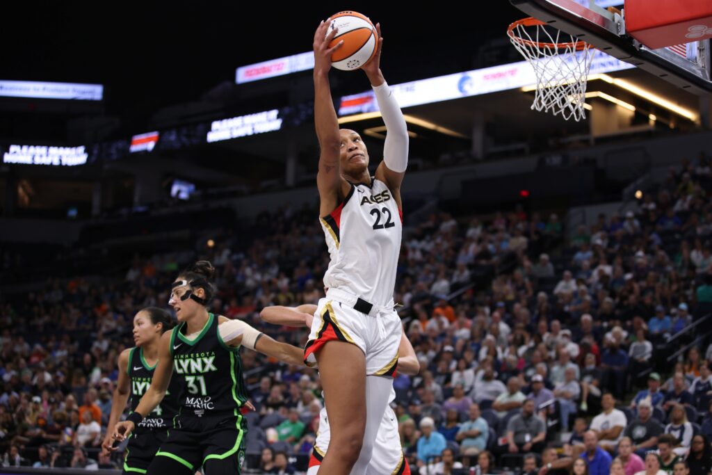 A’ja Wilson wearing her #22 Las Vegas Aces white uniform and jumping for a rebound with two Minnesota Lynx players in black and green jerseys behind her