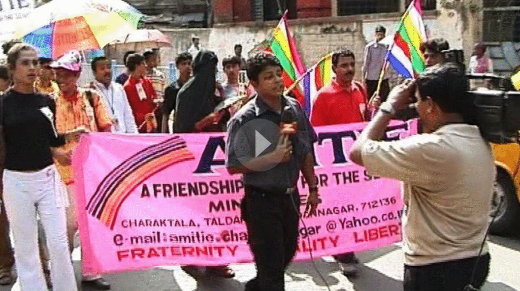 A Pride March in India with a reporter in front speaking to a news camera; behind the reporter march activists holding a pink banner and many others with rainbow flags and umbrellas.