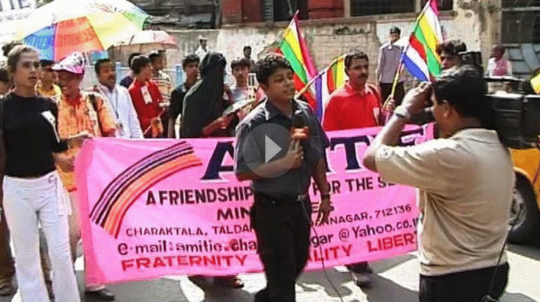 A Pride March in India with a reporter in front speaking to a news camera; behind the reporter march activists holding a pink banner and many others with rainbow flags and umbrellas.