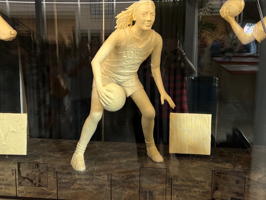 A life-size sculpture of Caitlin Clark dribbling a basketball, made entirely of butter, on display alongside other sculptures.