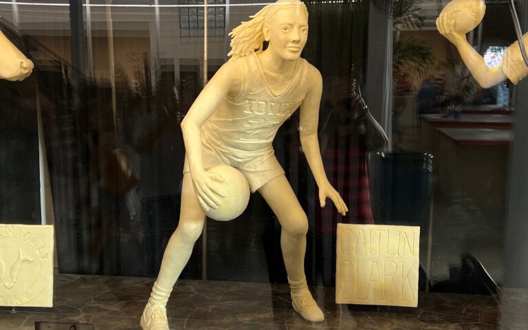 A life-size sculpture of Caitlin Clark dribbling a basketball, made entirely of butter, on display alongside other sculptures.