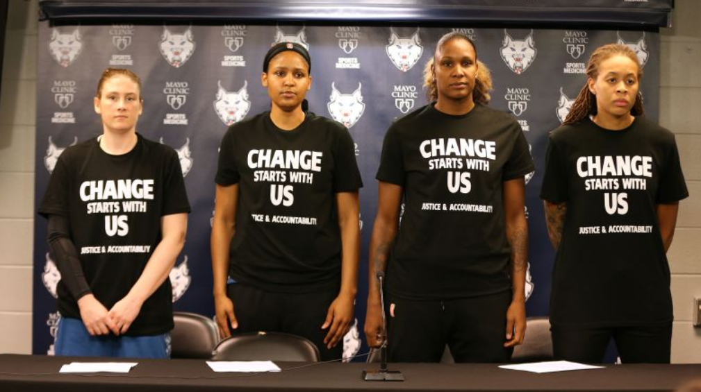 Four Minnesota Lynx players stand in front of a press conference table wearing black shirts that read "Change Starts With Us: Justice.& Accountability"