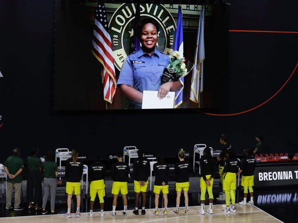 Seattle Storm players stand with their backs to the camera, facing the projection of a photo of Breonna Taylor. The players are wearing black warm-up shirts that say "Say Her Name" on the back