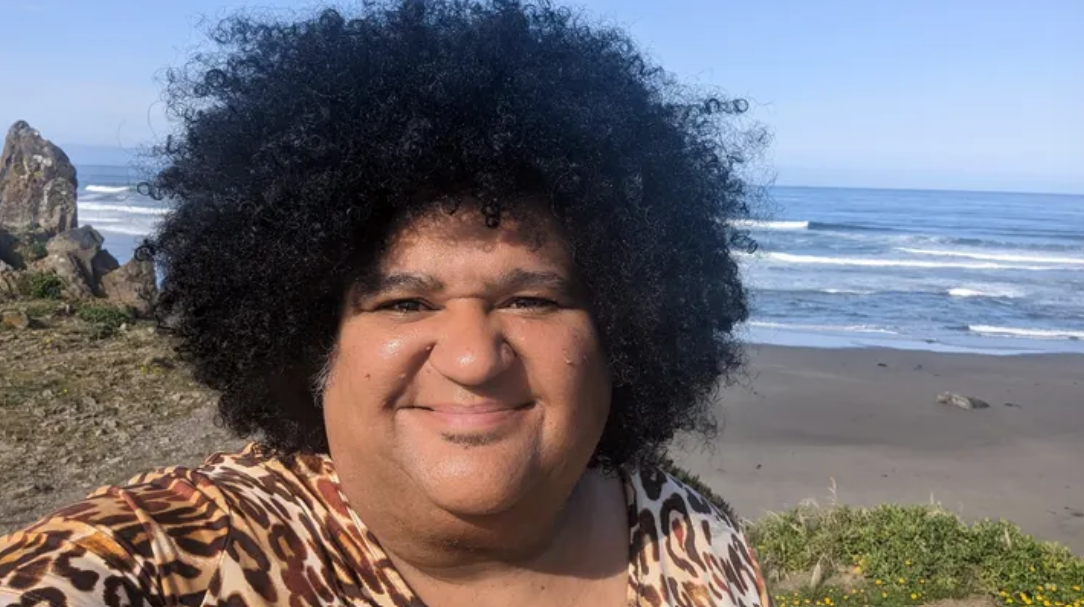Regent Brown: Selfie of a Black and Indigenous person with curly black hair smiling at the camera on a beach with a view of waves rolling in behind the person.